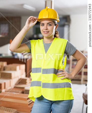 Confident female builder in yellow vest and hard hat at construction site 130772943