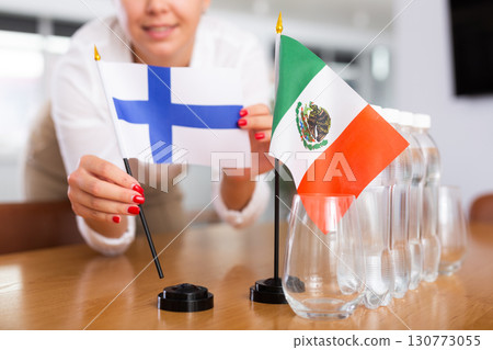 A woman in a white blouse with friendly smile arrangs flags of Mexico and Finland for meeting. 130773055