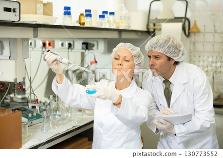 Middle-aged male and female researchers mixing reagents in test tube with lab pipette and writing report in laboratory 130773552