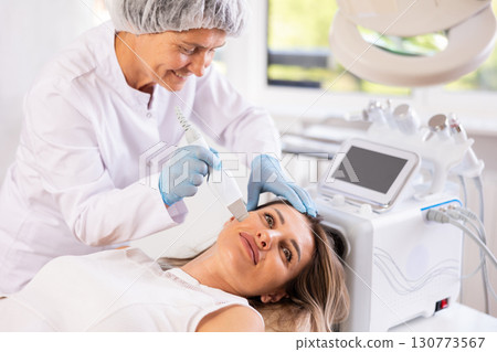 Young female client experiencing facial skin peeling with ultrasonic scrubber Young female client experiencing facial skin peeling with ultrasonic scrubber 130773567