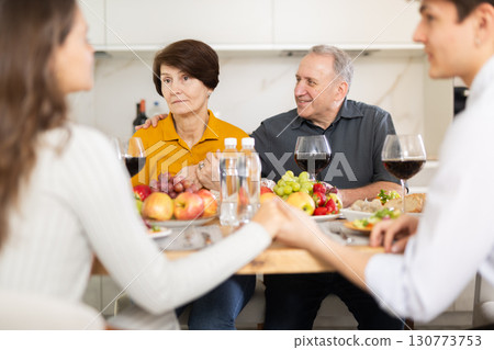 Puzzled elderly mother looking on with concern and surprise while adult son in love engaging in conversation with girlfriend at dinner table during family meal, young couple in love clasping hands Puzzled elderly mother looking on with concern and surprise while adult son in love engaging in conversation with girlfriend at dinner table during family meal, young couple in love clasping hands 130773753