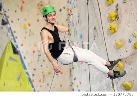 Sporty young guy in safety gear doing a difficult wall climb in climbing gym Sporty young guy in safety gear doing a difficult wall climb in climbing gym 130773793
