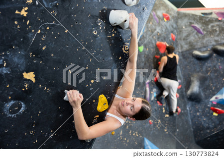 Sporty strong young female exercising in boulder climbing hall without rope, reaching new results, enjoying new challenges in amusement park 130773824