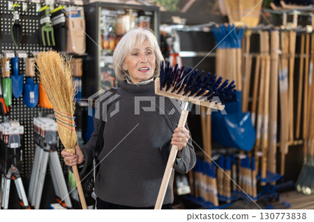 Senior woman in hardware store examines broom and besom. Senior woman in hardware store examines broom and besom. 130773838