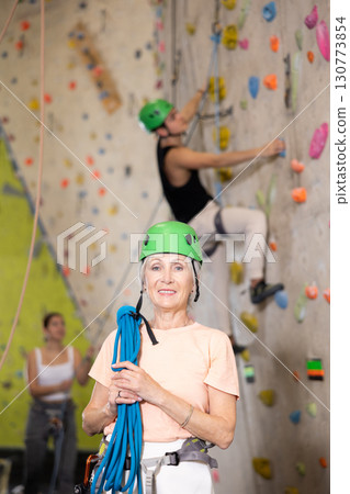 elderly positive slim woman with necessary equipment stands in gym near climbing wall 130773854