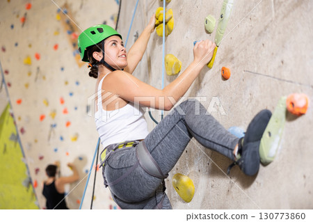 Sporty young girl in safety gear doing difficult wall climb in climbing gym Sporty young girl in safety gear doing difficult wall climb in climbing gym 130773860