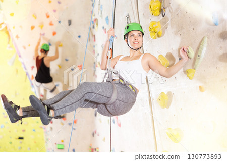 female climber athlete posing while hanging on her ropes near artificial climbing wall. female climber athlete posing while hanging on her ropes near artificial climbing wall. 130773893