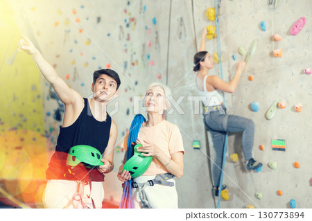 Young male trainer giving instructions to elderly woman about climbing artificial training rock wall in indoor bouldering gym 130773894