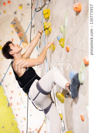 young positive man, insured by trainer, moving up artificial climbing wall indoors 130773957