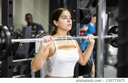 Caucasian woman doing exercises usining barbell and half rack in gym Caucasian woman doing exercises usining barbell and half rack in gym 130773975