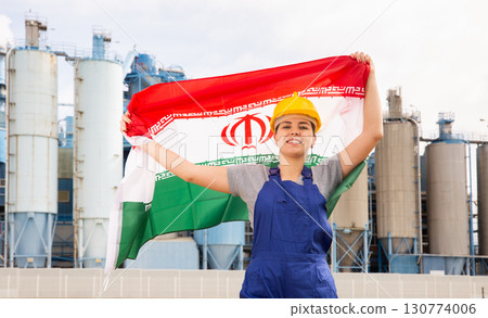 Happy girl in work clothes and hardhat with flag of iran standing in front of industrial scenery 130774006