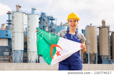 National flag of Algeria in the hands of girl in overalls against background of modern metallurgical plant National flag of Algeria in the hands of girl in overalls against background of modern metallurgical plant 130774017