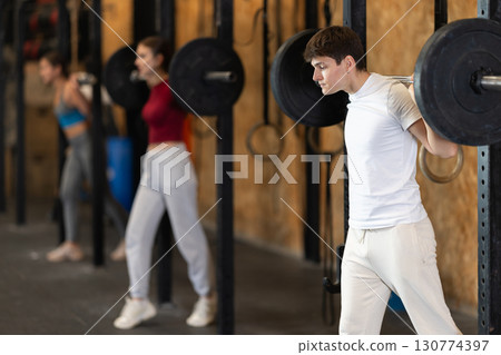 Slender guy trains muscles of arms and back using barbell and differentiated weighting in gym Slender guy trains muscles of arms and back using barbell and differentiated weighting in gym 130774397