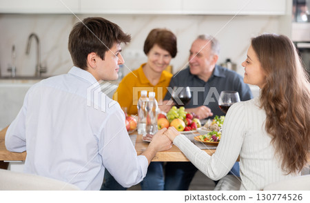 Couple sharing close moment at table, holding hands and locking eyes, while happy parents watching in background at family home. Blessing of parents 130774526