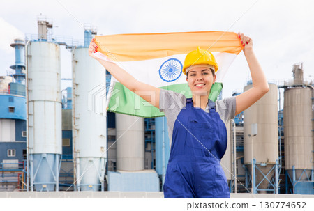 Young female engineer in helmet waving state flag of India while standing in front of big tanks at chemical plant 130774652