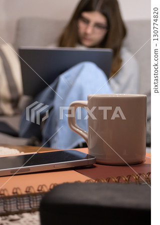 Cup of coffee and books in foreground and young female student in blurred background 130774820