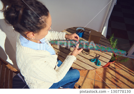 Woman Wrapping a Gift Outdoors on a Wooden Table with Festive Supplies Woman Wrapping a Gift Outdoors on a Wooden Table with Festive Supplies 130778130