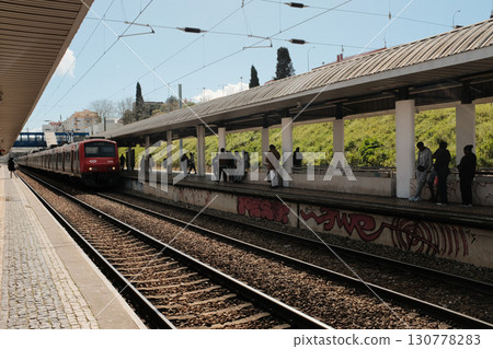 Red commuter train at suburban platform in Lisbon Portugal during sunny day with passengers waiting and graffiti on walls everyday public transportation scene in urban Europe. High quality photo 130778283