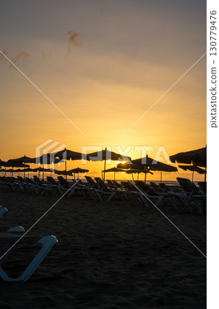 耀眼的朝陽從地平線升起,沙灘床和遮陽傘排列在富埃特文圖拉島的沙灘上 耀眼的朝陽從地平線升起,沙灘床和遮陽傘排列在富埃特文圖拉島的沙灘上 130779476