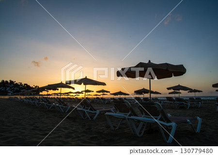 A dazzling morning sun rising from the horizon and beach beds and parasols lined up on the sandy coastline on Fuerteventura Island A dazzling morning sun rising from the horizon and beach beds and parasols lined up on the sandy coastline on Fuerteventura Island 130779480
