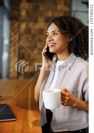 A Woman Relishing Her Coffee While Engaging in a Phone Conversation in a Cozy Cafe 130780022