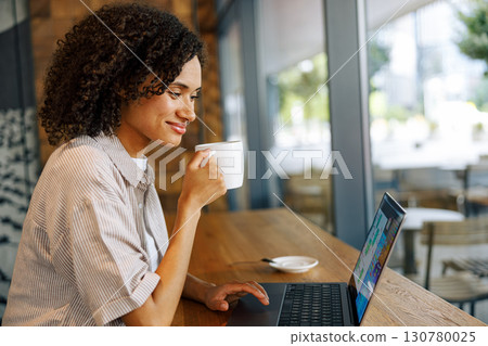 A Woman is Enjoying Her Coffee While Working on Her Laptop in a Modern Cafe Setting A Woman is Enjoying Her Coffee While Working on Her Laptop in a Modern Cafe Setting 130780025