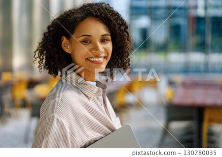 A cheerful, professional person smiling while working on a laptop in a lively urban setting 130780035