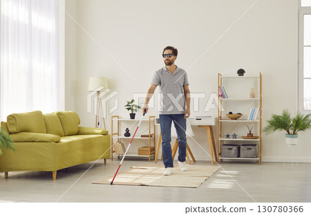 Bind young disabled man in casual clothes with cane walking in the living room at home. Bind young disabled man in casual clothes with cane walking in the living room at home. 130780366