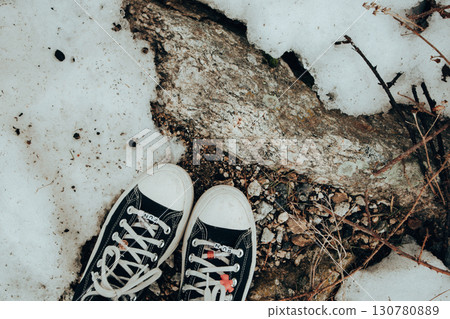 Black canvas sneakers on snow and rock, fun-themed shoelace charms, in a top view. A pair of black canvas sneakers 130780889