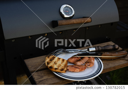 A delicious grilled meal featuring sausages and toast is ready for the summer season. A barbecue grill with food on a wooden surface, ready to be served. The scene is set outdoors on a sunny day. A delicious grilled meal featuring sausages and toast is ready for the summer season. A barbecue grill with food on a wooden surface, ready to be served. The scene is set outdoors on a sunny day. 130782015