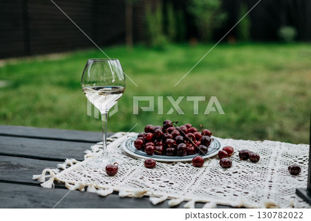 A glass of white wine and a plate of raw fresh red cherries on a wooden terrace. Summertime picnic outdoors. No people. Rustic still life. 130782022