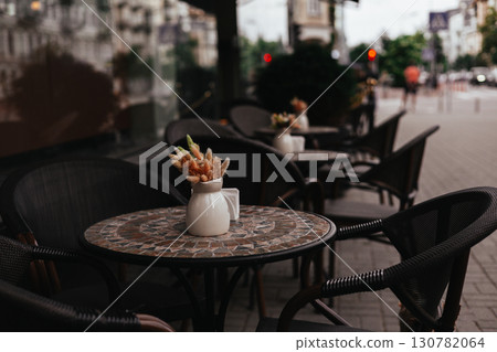 A charming cafe terrace with tables, chairs, and a vase of dried flowers adds a touch of charm awaits customers. An inviting outdoor cafe scene. 130782064