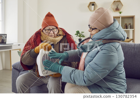 Senior family couple in winter clothes warming up by electric fan heater at home 130782354