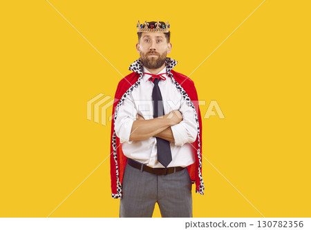 Portrait of young confident man in suit with crown looking at camera on yellow background. 130782356