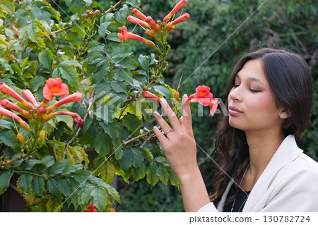 Young Indian woman with long dark hair standing near orange blossoms, gently raising her hand toward the flowers. A lifestyle and cultural beauty portrait Young Indian woman with long dark hair standing near orange blossoms, gently raising her hand toward the flowers. A lifestyle and cultural beauty portrait 130782724