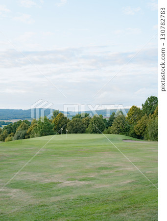 Expansive golf course with vibrant green grass, surrounded by lush trees under a partly cloudy sky. Two flags indicate the holes, creating a serene and tranquil setting 130782783