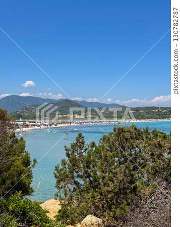 Sunny summer day at a popular beach in Sardinia with turquoise waters, sunbathers, and distant green mountains. 130782787