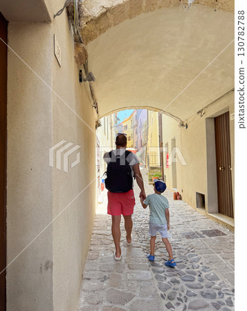 A father and son walk hand in hand under a stone archway in a charming narrow alley of a Mediterranean old town on a sunny summer day. A father and son walk hand in hand under a stone archway in a charming narrow alley of a Mediterranean old town on a sunny summer day. 130782788