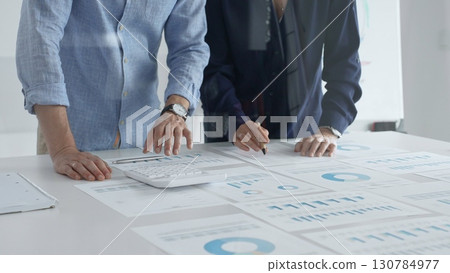 Business professionals analyzing financial charts, gesturing over spreadsheets during collaborative meeting in contemporary workspace, discussing data driven insights 130784977