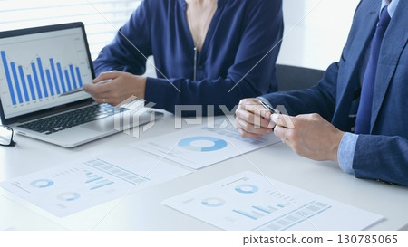 Businessman and businesswoman are analyzing financial data on a laptop and printed documents, pointing at charts and graphs during a meeting in bright office. Business people and finance concept 130785065