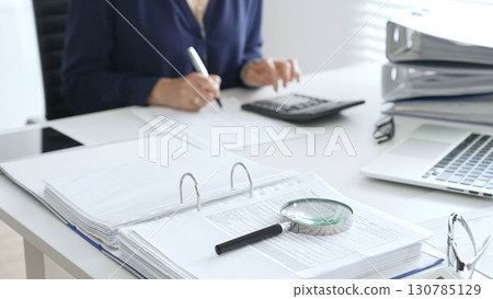 Professional analyzing financial data, writing notes near laptop, magnifying glass, documents, binders on workplace desk. Audit and taxes concept Professional analyzing financial data, writing notes near laptop, magnifying glass, documents, binders on workplace desk. Audit and taxes concept 130785129