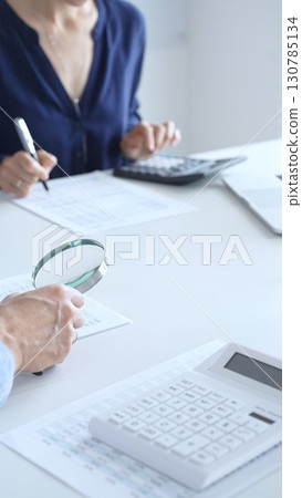 Two accountants analyzing financial reports with magnifying glass and documents, performing accounting and auditing tasks in the office, close up of hands. Audit and taxes in business Two accountants analyzing financial reports with magnifying glass and documents, performing accounting and auditing tasks in the office, close up of hands. Audit and taxes in business 130785134