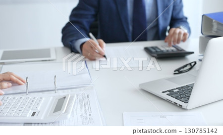 Two accountants are collaborating on a project, using a calculator and taking notes, surrounded by documents and technology in a busy office environment. Audit and taxes in business Two accountants are collaborating on a project, using a calculator and taking notes, surrounded by documents and technology in a busy office environment. Audit and taxes in business 130785142