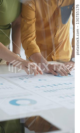 Professional team collaborating, reviewing financial documents and graphs on laptop with printed data spread across conference table in contemporary workspace. Business people concept Professional team collaborating, reviewing financial documents and graphs on laptop with printed data spread across conference table in contemporary workspace. Business people concept 130785176