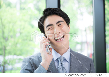 A smiling business man in a suit making a phone call by a window with fresh greenery, looking at the camera, making an inquiry or application A smiling business man in a suit making a phone call by a window with fresh greenery, looking at the camera, making an inquiry or application 130786493