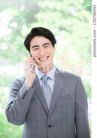 一位穿著西裝、微笑的商人在綠樹成蔭的窗邊打電話，看著鏡頭，進行詢問或申請 130786495