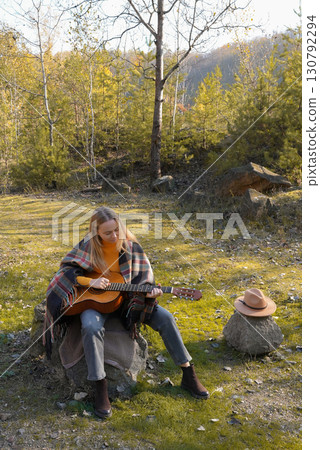 Blonde woman playing string guitar outdoors in autumn forest. Concept of sound therapy, mental health and wellness rituals. Calmness tranquility audio-sensory practices. Aura farming energy 130792294