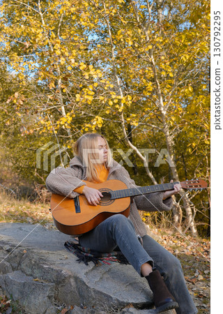 Beautiful blonde girl playing acoustic guitar and singing relaxing enjoying outdoors on sunny autumn day. Hippie woman in boho style learning and practicing playing musical instrument. Fall leaves 130792295