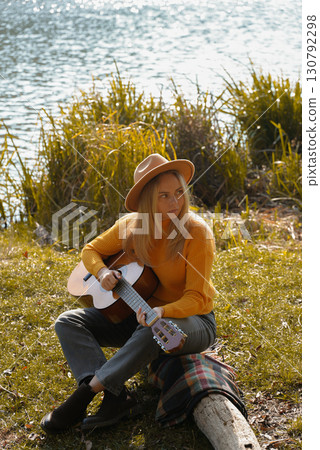 Beautiful blonde girl playing acoustic guitar and singing relaxing enjoying outdoors on sunny autumn day. Hippie woman in boho style learning and practicing playing musical instrument. Fall leaves 130792298