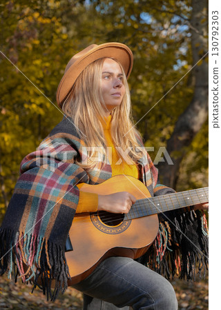 Blonde woman playing string guitar outdoors in autumn forest. Concept of sound therapy, mental health and wellness rituals. Calmness tranquility audio-sensory practices. Aura farming energy 130792303